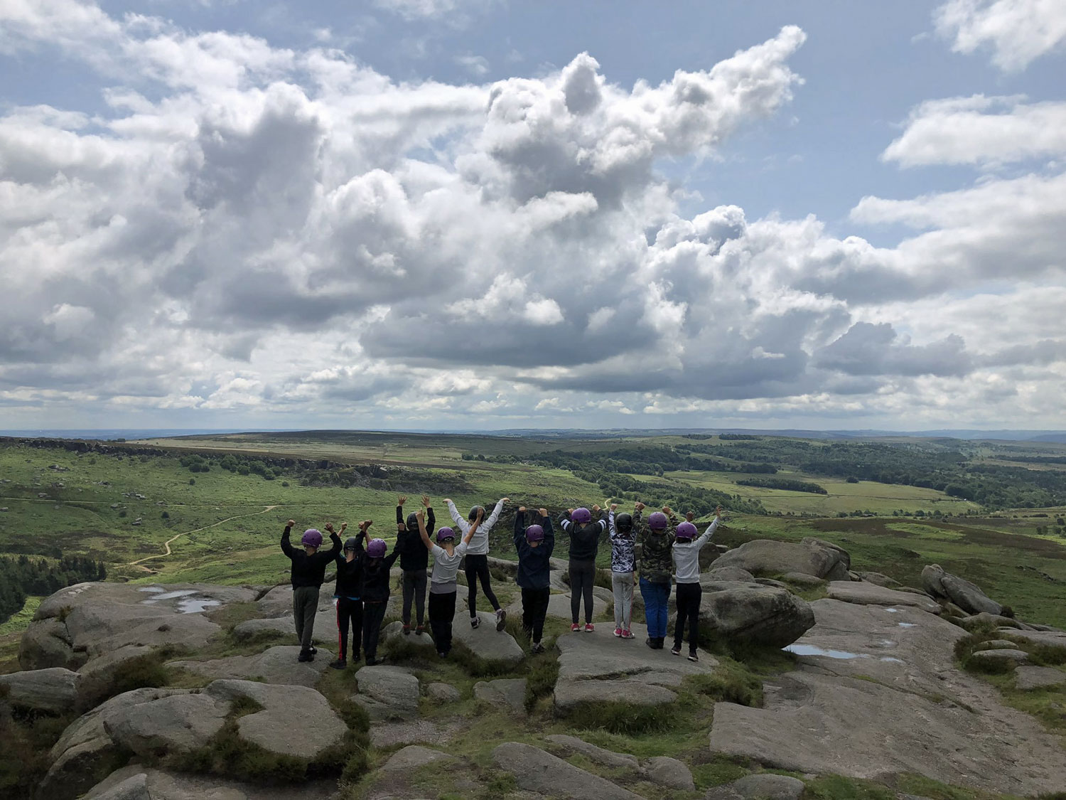 School children in helmets stand on on an edge looking out over the vast Peak District scenery with their arms raised jubilantly