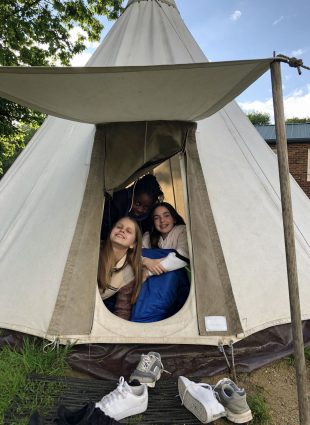 Children smile from within a teepee as they peek out from beneath the entrance flap