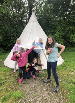 Children give the camera a thumbs up as they pose in front of their teepee