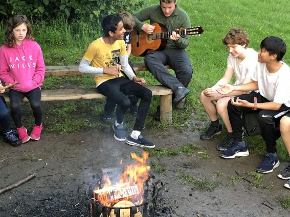 Young adults sit around the campfire one play the guitar and another sings