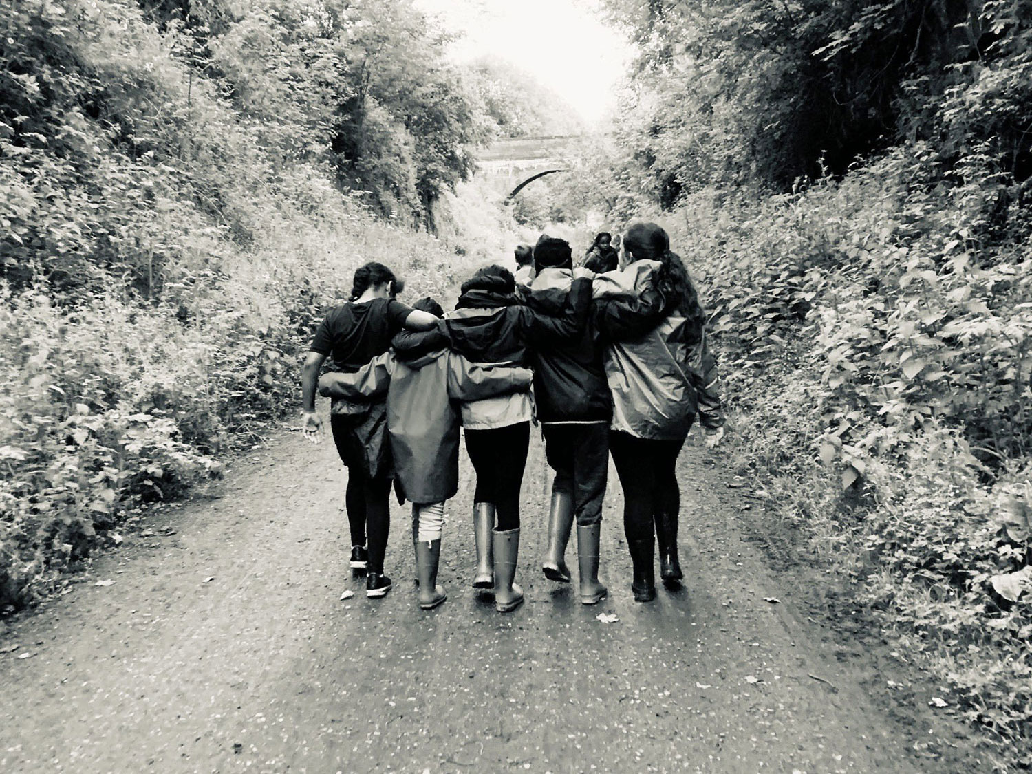 Black and white photo taken from behind shows a group of five girls walking down the Monsal Trail with their arms around eachother