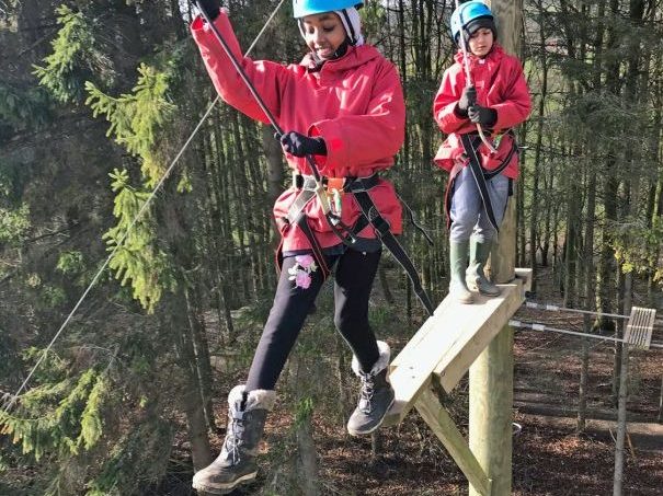 One student bravely takes the leap of faith at the top of the Sky Ropes, while another child looks on in fear