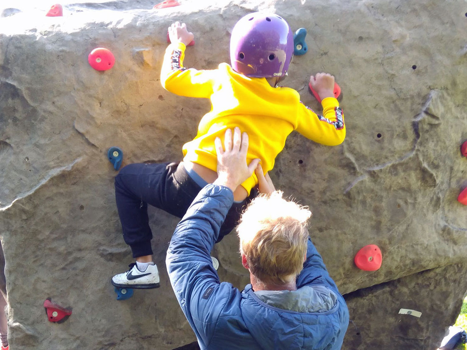 The instructor supports a small child who is climbing the outdoor boulder