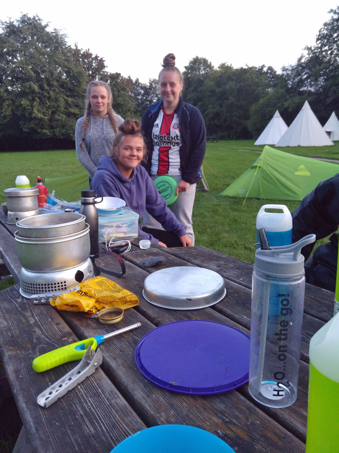 D of E students stand by a picnic bench out in the fields with their tent assembled in the background