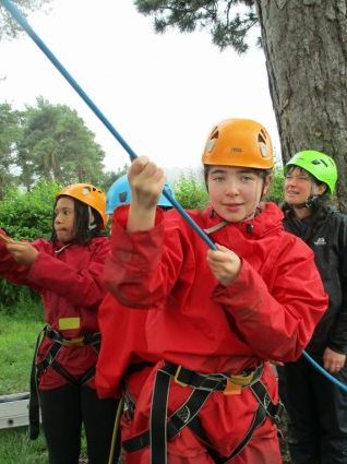 School children hold the belaying ropes and look up from the sidelines while their friends climb the cargo net