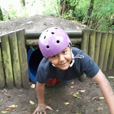 Boy crawls out of tunnel in the woods of Thornbridge Outdoors with a helmet on, he's muddy and smiling