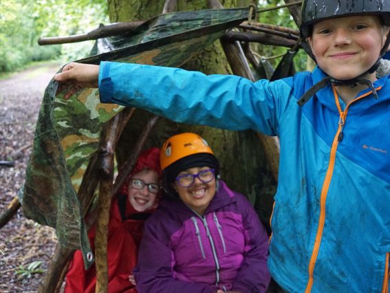 Boy holds up tarpaulin sheet on their den in the woods and we can see two classmates smiling as they sit inside the den