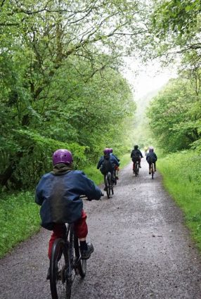 Photo taken from behind shows school children riding bikes along a green bushy stretch of the Monsal Trail